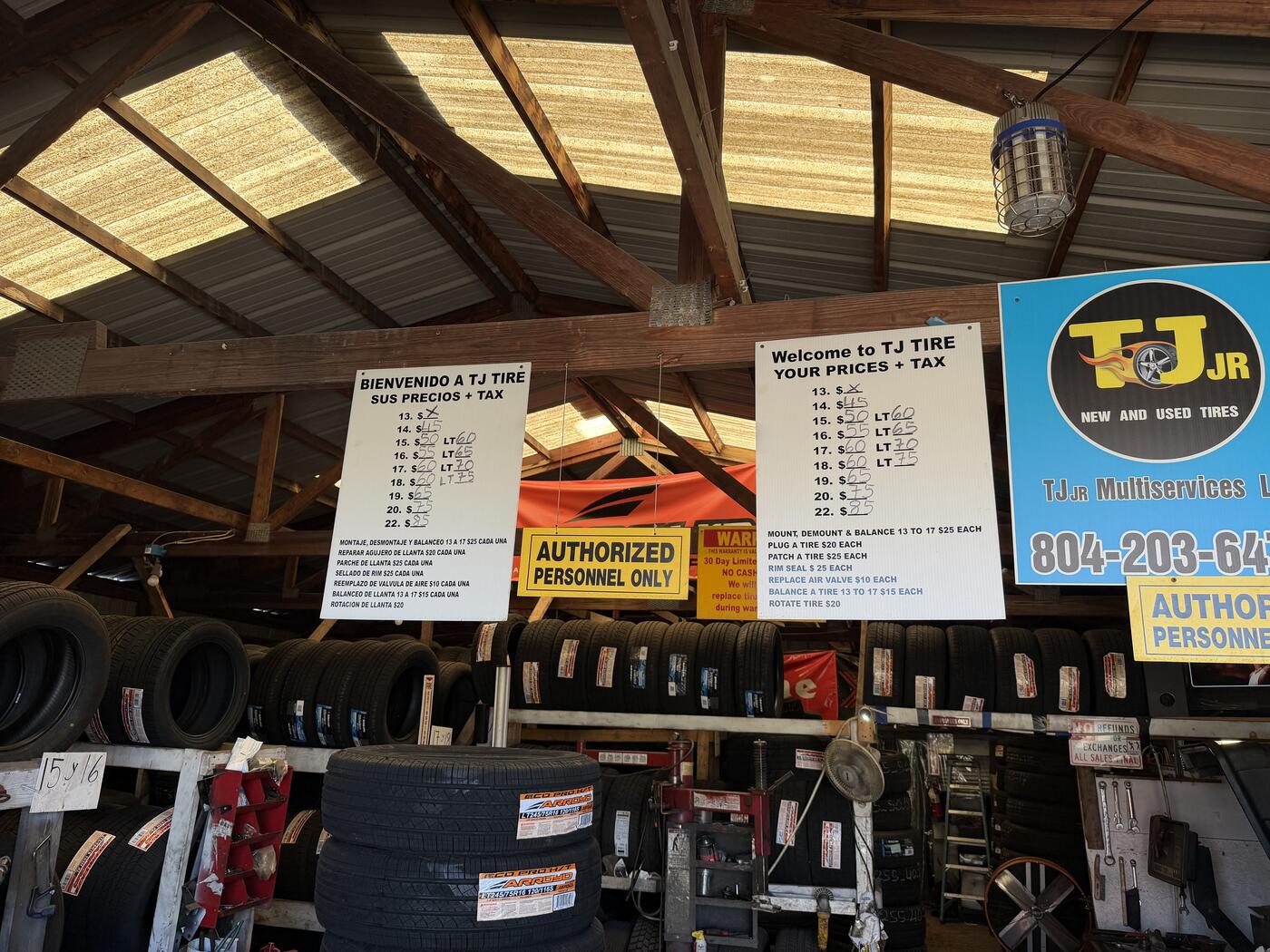 The price boards hanging inside TJ Jr under the wood-beam roof — 'Welcome to TJ Tire / Bienvenido a TJ Tire — Your Prices + Tax' with tire sizes and the TJ Jr logo